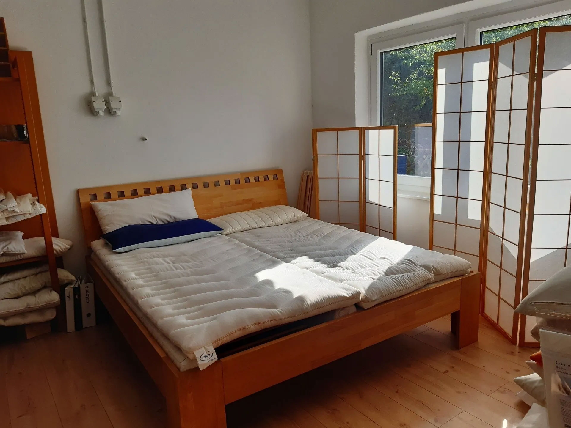 Probeliegeraum mit speltex Naturprodukten. Bett aus Holz mit speltex-Matratzen und -toppern sowie verschiedenen Kissen. Ansicht vom Fußende des Bettes aus. Links im Regal gibt es weitere Kissen und Produkte zu sehen. Rechts neben dem Bett steht ein Raumtrenner aus hellem Holz und transparenten Scheiben vor dem Fenster.